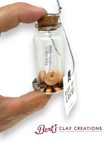 Glass bottle with cork lid containing a small fortune cookie, held by a hand against a white background. 'Bert's Clay Creations' branding visible.